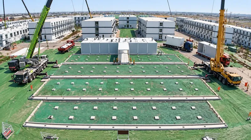 Cranes setting modules beside rows of prepared foundation pads, with finished two-story modular buildings behind
