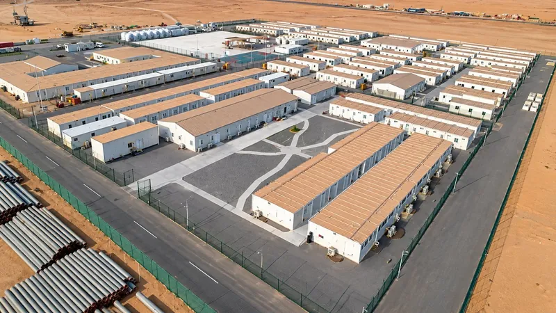 Aerial view of a fenced desert camp of long single-story modular buildings around a paved courtyard