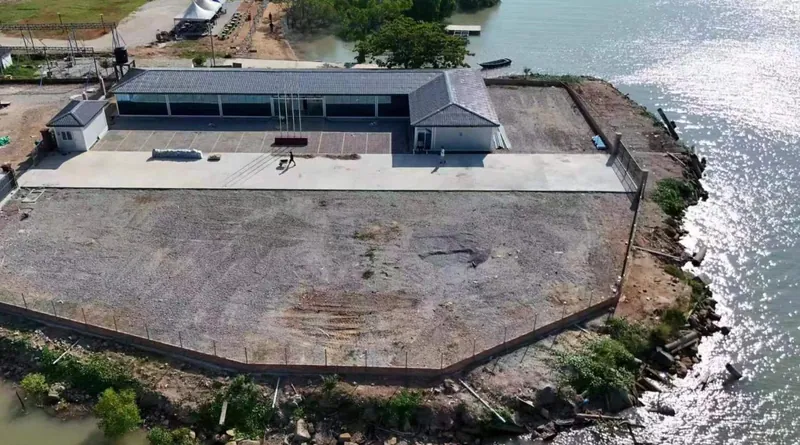 Aerial view of a long gray-roofed building on a paved plot at the water’s edge
